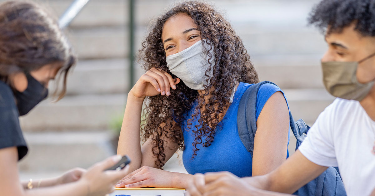 três jovens estudantes, com máscaras de proteção, conversam em volta de uma mesa.