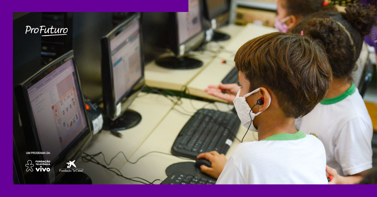 Imagem mostra dois alunos utilizando computadores. Eles estão usando uniforme escolar e máscara.