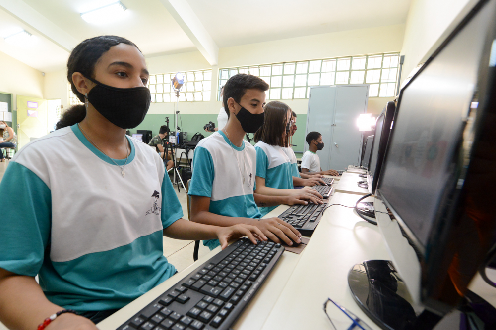 Imagem mostra vários alunos em uma sala de aula. Todos estão utilizando computadores. Todos usam máscara e uniforme da escola