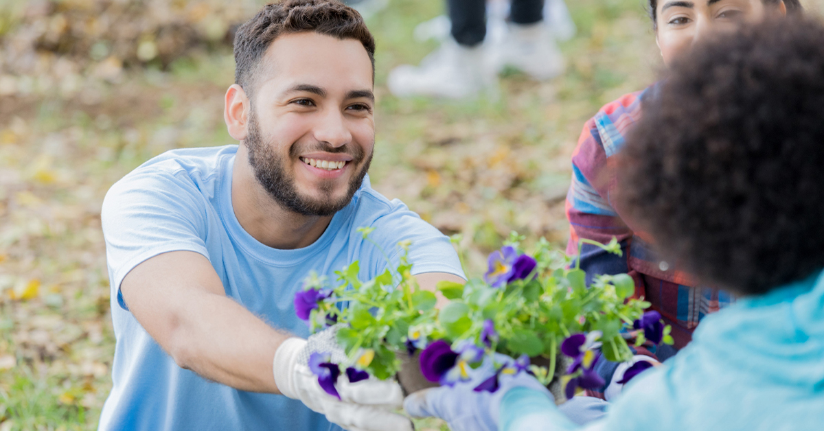 Imagem mostra três jovens em um local que parece ser um parque. Um dos jovens está segurando um vaso de flores e entregando para uma das jovens. A terceira jovem observa os dois jovens interagindo.