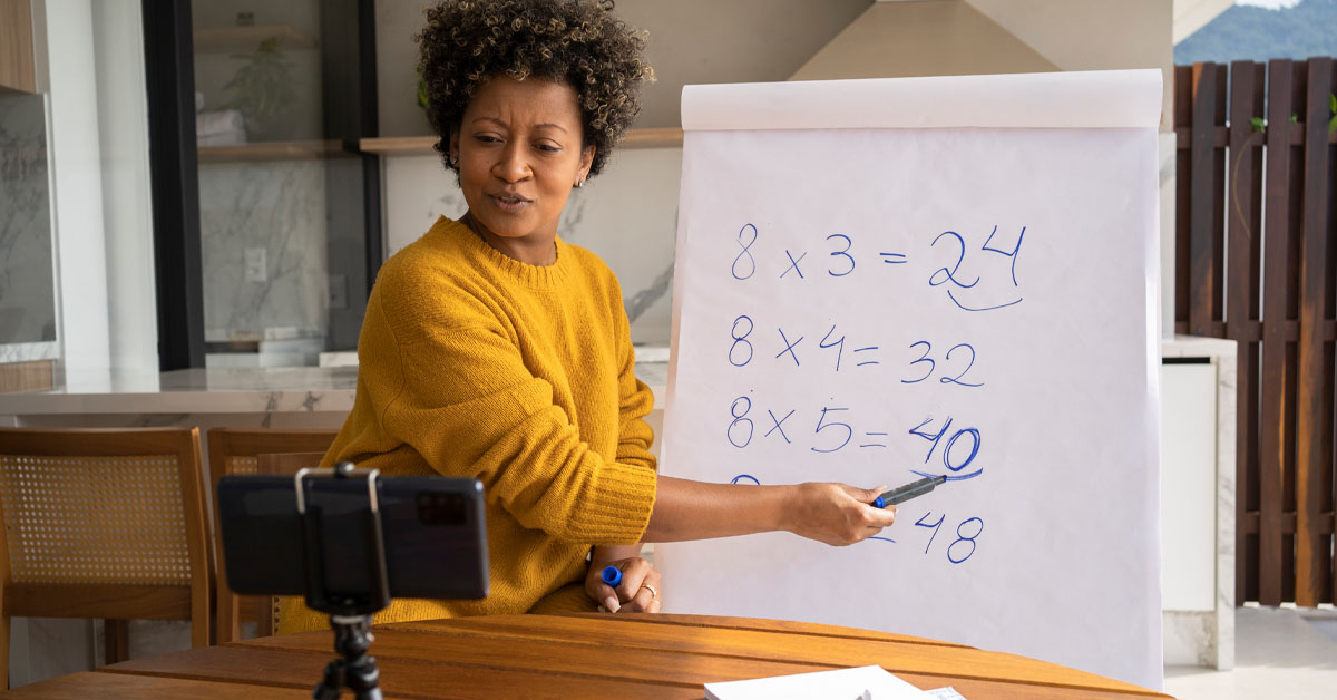 Imagem mostra uma educadora ensinando matemática. Ela está apontando para alguns números apresentados em um painel. À sua frente há um celular, o que nos faz entender que a aula que ela está dando é on-line.