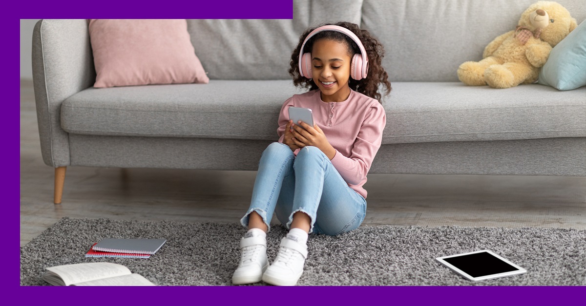 Imagem mostra uma menina em uma sala de estar. Ela está sentada sobre um tapete, encostada em uma poltrona, está usando fones de ouvidos e segurando um aparelho que parece ser um celular.