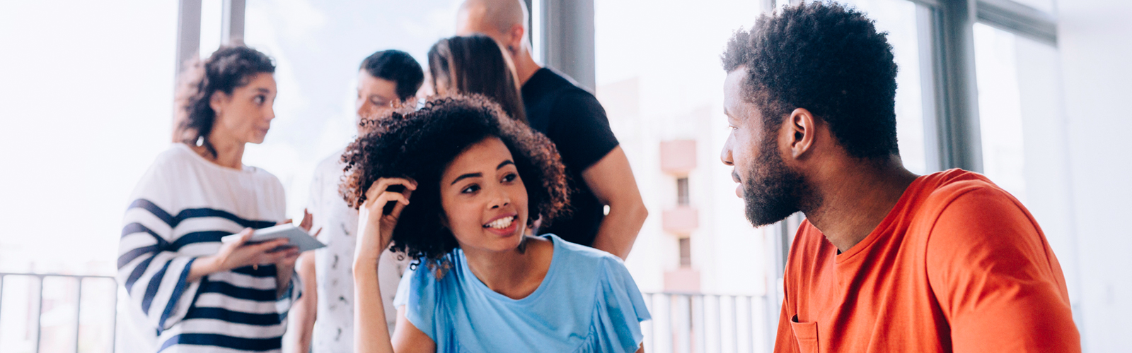 Uma jovem negra de camiseta azul conversa com um jovem negro de camiseta laranja em uma sala. Ao fundo estão quatro pessoas de pé, conversando entre elas.