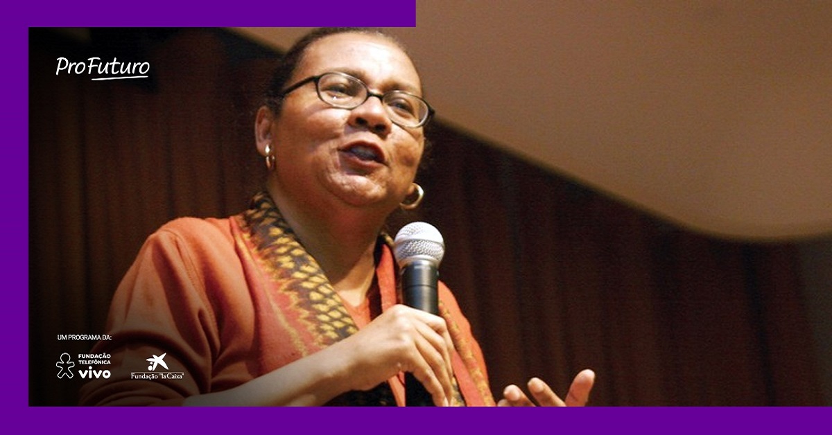 Imagem mostra a ativista bell hooks. Ela está segurando um microfone e parece estar palestrando. Usa óculos, brincos dourados e uma blusa vermelha.