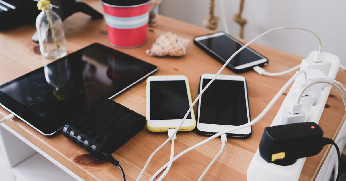 Imagem mostra uma mesa com diversos aparelhos eletrônicos como celulares, tablet e a o monitor de um computador. Os três aparelhos de celular estão conectados a cabos e estão sendo carregados em tomadas