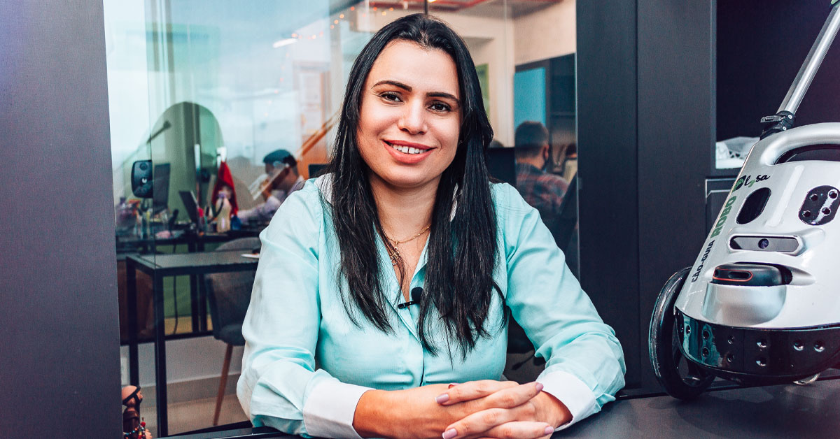 Imagem mostra a professora Neide Sellin, criadora do cão-guia robô. Ela usa uma camisa azul, tem cabelo comprido, está sorrindo para a foto e com os braços apoiados em uma mesa. Ao seu lado, é possível ver o robô. Ao fundo, é possível ver algumas pessoas trabalhando em um ambiente que parece ser um escritório.