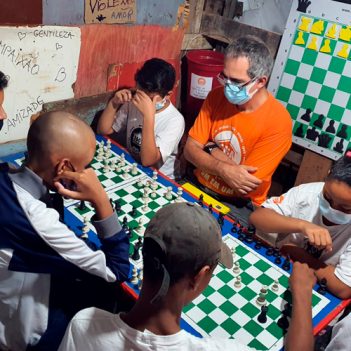 Imagem mostra alunos do projeto Casa Xadrez em volta de uma mesa onde há três tabuleiros do jogo. Eles estão acompanhados de um adulto, que parece ser o professor, e estão concentrados observando as peças do jogo