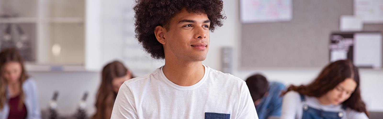 Em uma sala de aula, um estudante negro, de camiseta branca, sorri enquanto olha para o lado, pensativo. Ao fundo, estão quatro jovens estudando, sentados.