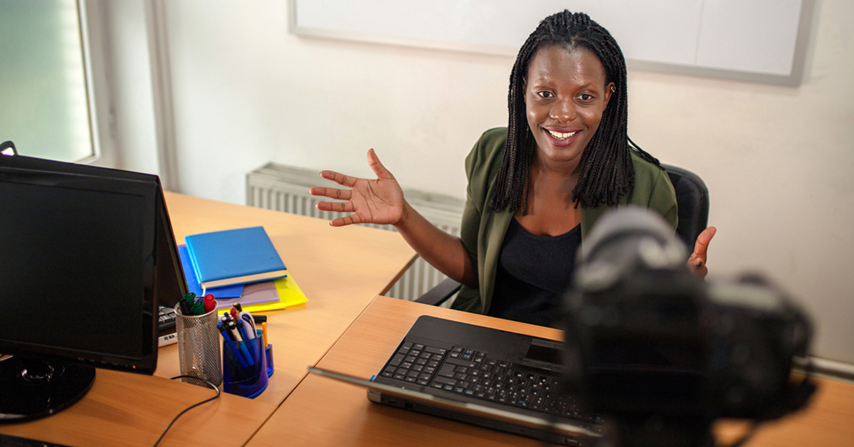 Imagem mostra uma educadora em uma sala de aula. Ela está sentada em frente a uma câmera. Parece estar dando aula on-line. Sobre a mesa, há um notebook, cadernos e canetas. A professora é negra e tem cabelos compridos trançados. Usa uma blusa preta e uma camisa verde por cima.