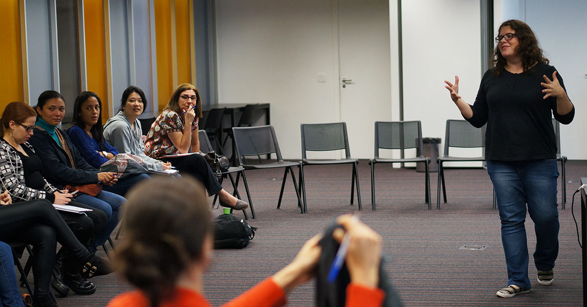 Imagem mostra uma sala de aula onde é possível ver alunas sentadas assistindo à aula de uma professora, que está em pé, conversando com as alunas.