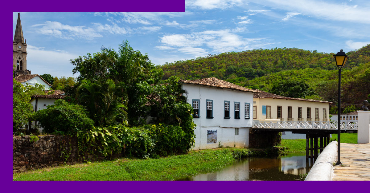 Imagem mostra um casarão antigo, cercado por árvores e um riacho. Ao fundo, é possível ver a torre de uma igreja