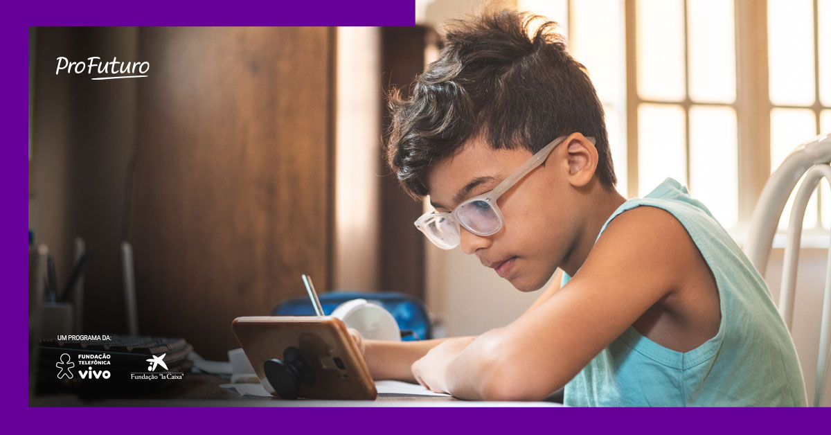 Imagem mostra um menino estudando com o apoio de um celular. Ele usa óculos, está com um lápis na mão direita, escrevendo em um caderno. O celular e o caderno estão sobre uma mesa.