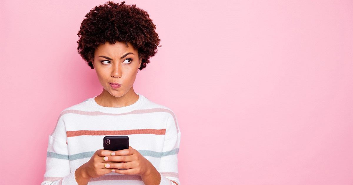 Imagem mostra uma jovem negra segurando um celular. Ela está com um semblante de dúvida. Usa blusa branca com listras nas cores azul e rosa.