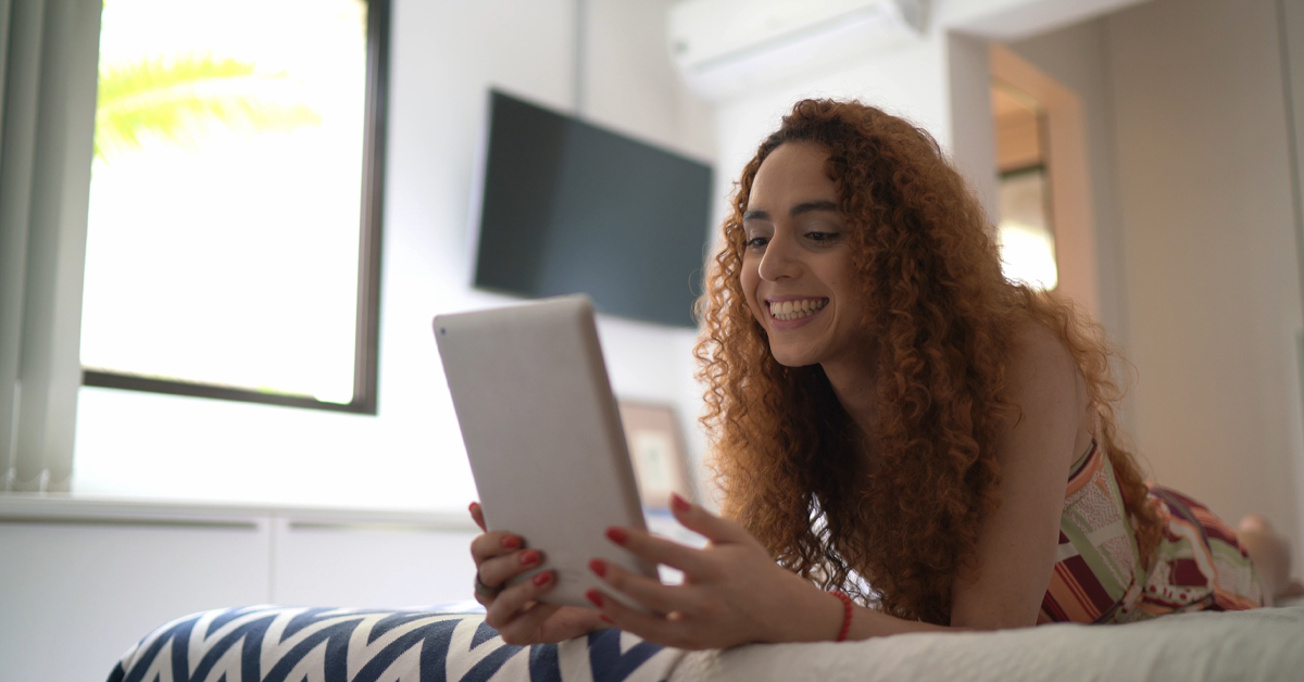 Imagem mostra uma jovem branca, de cabelos encaracolados e compridos, deitada sobre uma cama. Ela está segurando um tablet. No ambiente, é possível ver ao fundo uma televisão na parede