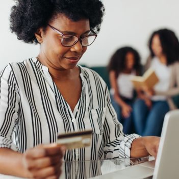 Imagem mostra uma mulher negra utilizando um notebook. Ela segura um cartão de crédito na mão direita, o que dá a entender que ela está fazendo uma transação financeira pela internet. Ao fundo, é possível ver uma mulher e uma menina sentadas em um sofá.