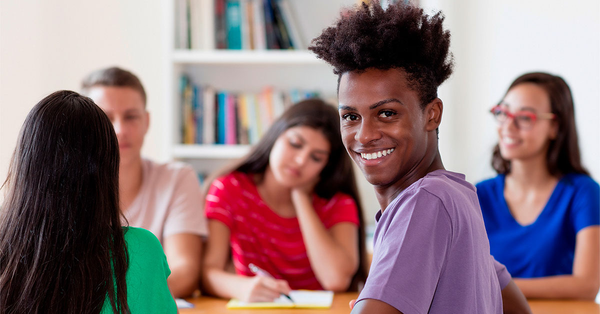 Imagem mostra cinco jovens reunidos, sentados ao redor de uma mesa, conversando. São dois rapazes e três moças. Um dos jovens é negro e está sorrindo.