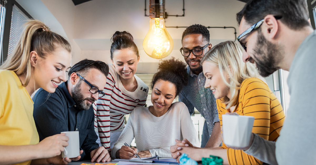 imagem mostra sete jovens empreendedores reunidos. São quatro mulheres e três homens. eles estão em volta de uma mesa. Uma das jovens está anotando algo em um caderno e mostrando para os demais jovens.