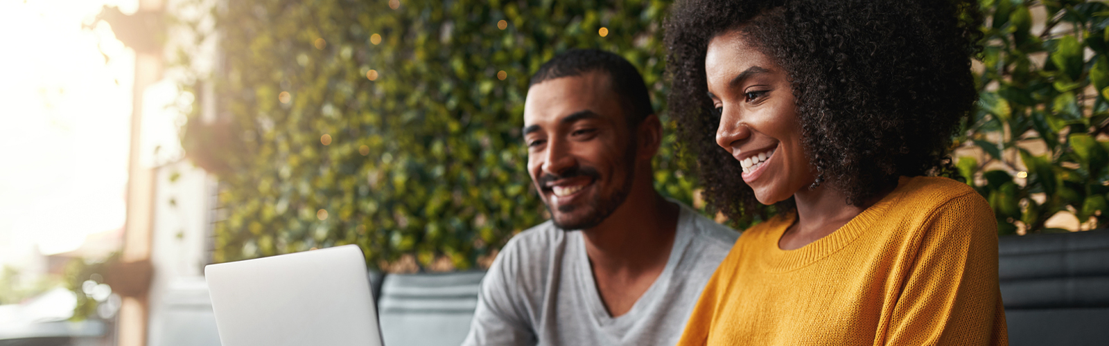 Em foto ambientada com árvores em um lugar aberto duas pessoas sorriem enquanto olham para a tela de um computador. O jovem, é um rapaz negro e veste camiseta cinza e a moça ao lado dele é negra, de cabelos crespos e usa um suéter laranja.