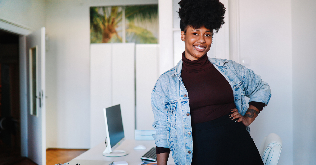 Imagem mostra uma mulher negra empreendedora. Ela usa blusa preta e jaqueta jeans, está com a mão esquerda na cintura. É possível ver um notebook em cima de uma mesa na sala onde a mulher está