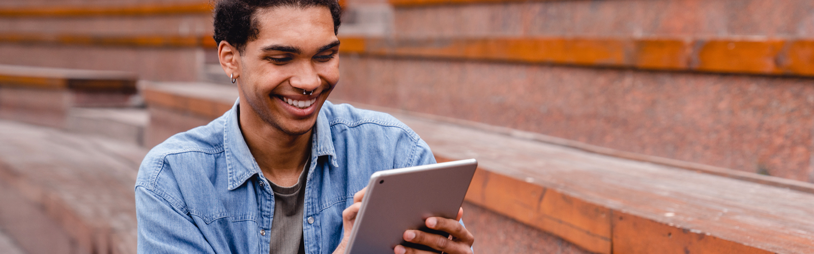 Um jovem está sentado sozinho em uma arquibancada sorrindo enquanto mexe em um tablet. Ele é negro, tem um piercing no nariz e veste uma camisa azul jeans e uma camiseta cinza por baixo.