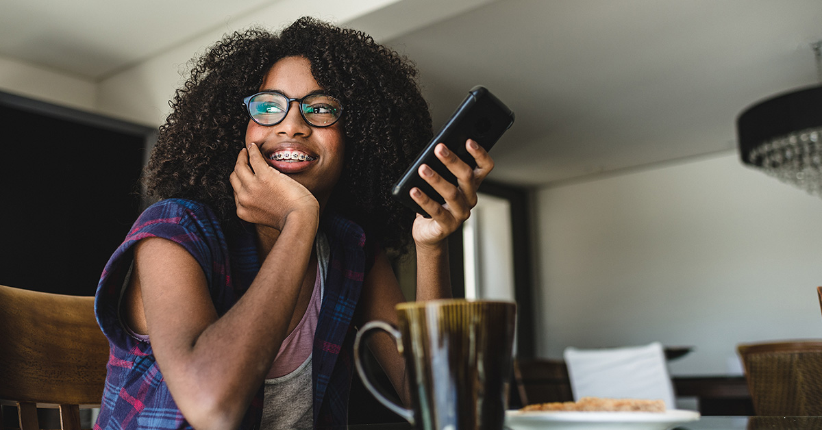 Menina de óculos e cabelos cacheados sorri com o rosto apoiado em uma das mãos e segura um smartphone na outra. Imagem ilustra pauta sobre o Pense Grande Digital, aplicativo que oferece formação gratuita em empreendedorismo social.