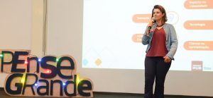 Mila Gonçalves, gerente de programas sociais da Fundação Telefônica Vivo, está de jaqueta jeans falando ao microfone em cima do palco do Demoday do Pense Grande, evento que premia jovens empreendedores.
