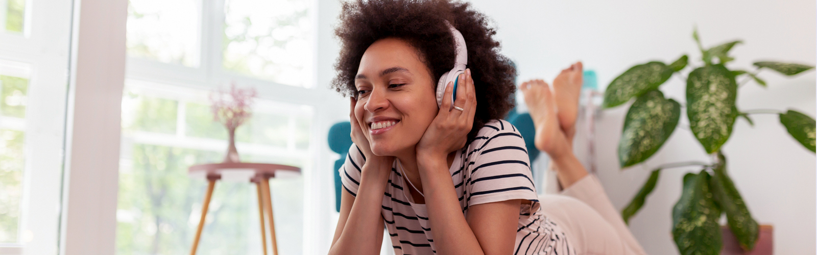 Uma jovem negra está em uma sala sorrindo e com fones de ouvido na cabeça escutando um podcast. Ao mesmo tempo que sorri, segura os fones com as mãos.