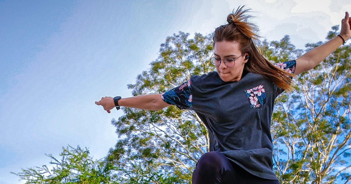 Imagem mostra a jovem skatista Isa Corleone. Ela está fazendo uma manobra aérea com o skate em m local que parece ser um parque. Usa calça e camiseta na cor azul escura. Ela tem cabelos longos e usa óculos.