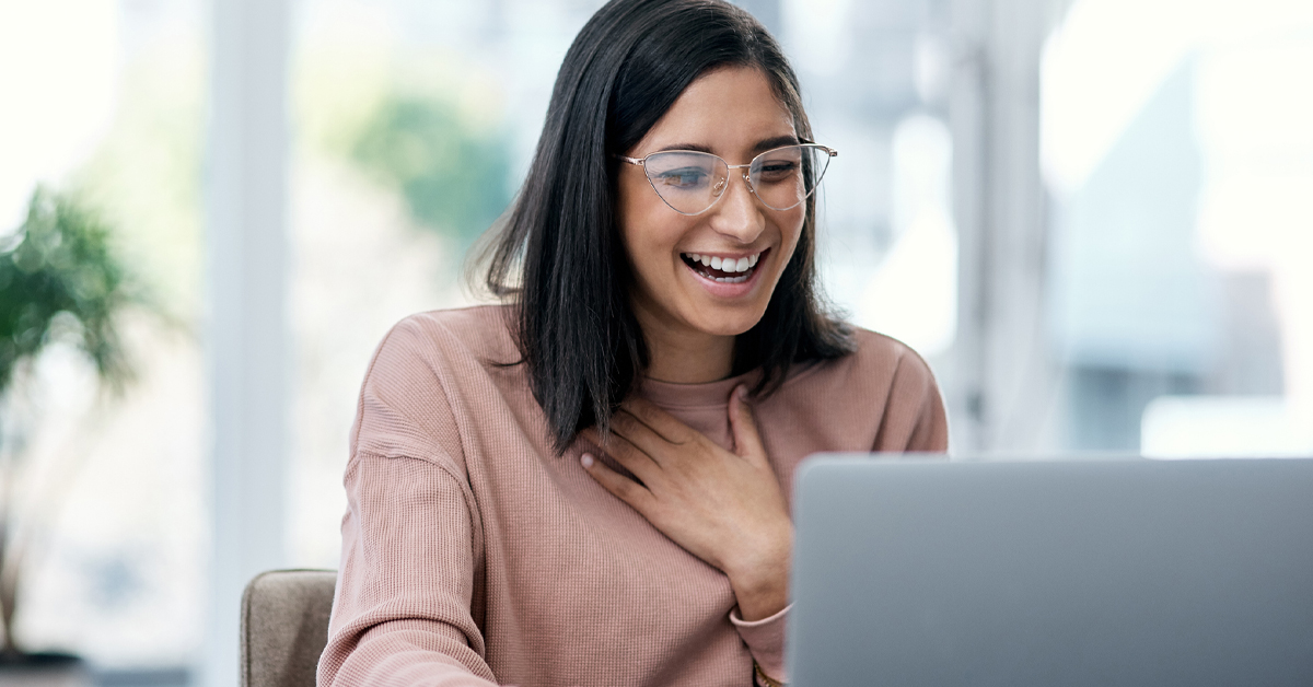 Imagem mostra uma jovem sorrindo. Ela está em frente a um computador. Ela usa óculos e uma blusa na cor rosa.