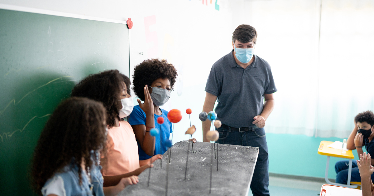 Imagem mostra uma sala de aula. Três alunos estão à frente da classe, apresentando um trabalho com uma maquete de planetas. Ao lado, o professor observa a apresentação, assim como demais alunos da classe. Todos usam máscara de proteção.