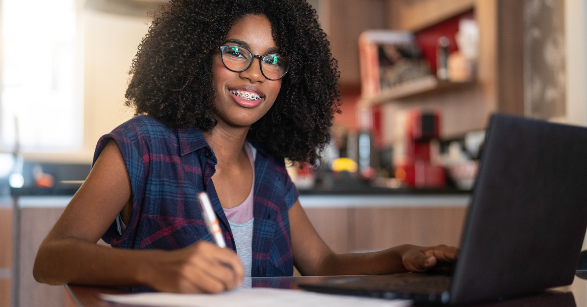 Imagem mostra uma jovem negra em um ambiente que parece ser a sua casa. Ela está sentada, em uma mesa à sua frente há um notebook preto e com a mão direita ela segura uma caneta. Na mesa, também há papéis. A jovem usa óculos, uma blusa branca e um colete xadrez, e sorri para a foto.