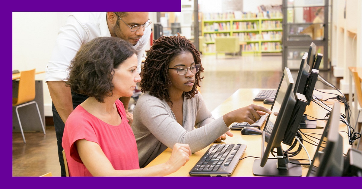 Imagem mostra duas professoras sentadas em frente a um computador, uma delas é negra, e um professor, que está em pé, atrás das professoras. Os três parecem estar conversando e analisando algo que está sendo mostrado no computador. Provavelmente um conteúdo relacionado ao desenvolvimento de competências digitais