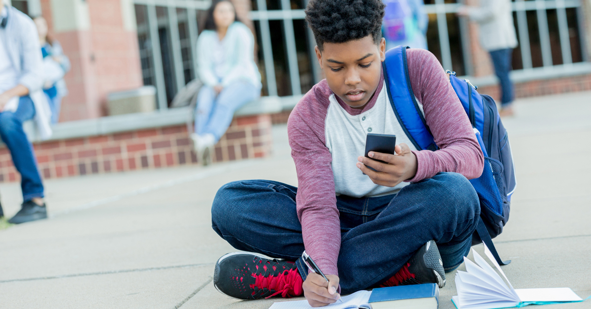 Imagem mostra um estudante negro, sentado no chão de uma área aberta em uma escola. Ele faz anotações em um caderno, enquanto consulta método de ensino de matemática em seu celular. Ao fundo, é possível ver outros estudantes no ambiente.