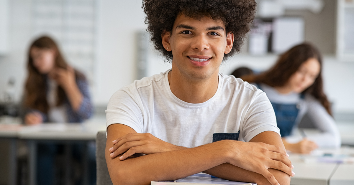 Imagem mostra um jovem negro em sala de aula. Ele sorri para a foto. Usa uma camiseta branca. Está sentado em uma carteira escolar e está com os braços cruzados. Ao fundo, é possível ver outros estudantes.