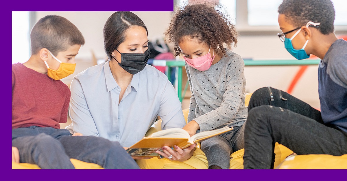 Imagem mostra uma educadora lendo um livro para três crianças, sendo uma menina e um menino negros e um menino branco. Todos usam máscara de proteção. A menina está ao lado da educadora apontando para o livro.