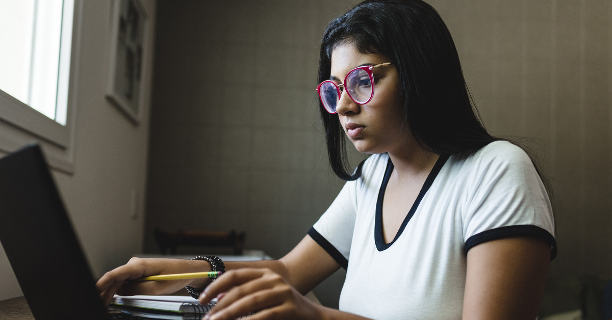 Imagem mostra uma jovem utilizando um notebook preto, que está sobre uma mesa. Ao lado, há um caderno. A jovem segura na mão direita um lápis. Ela está usando uma camiseta branca e óculos rosa.