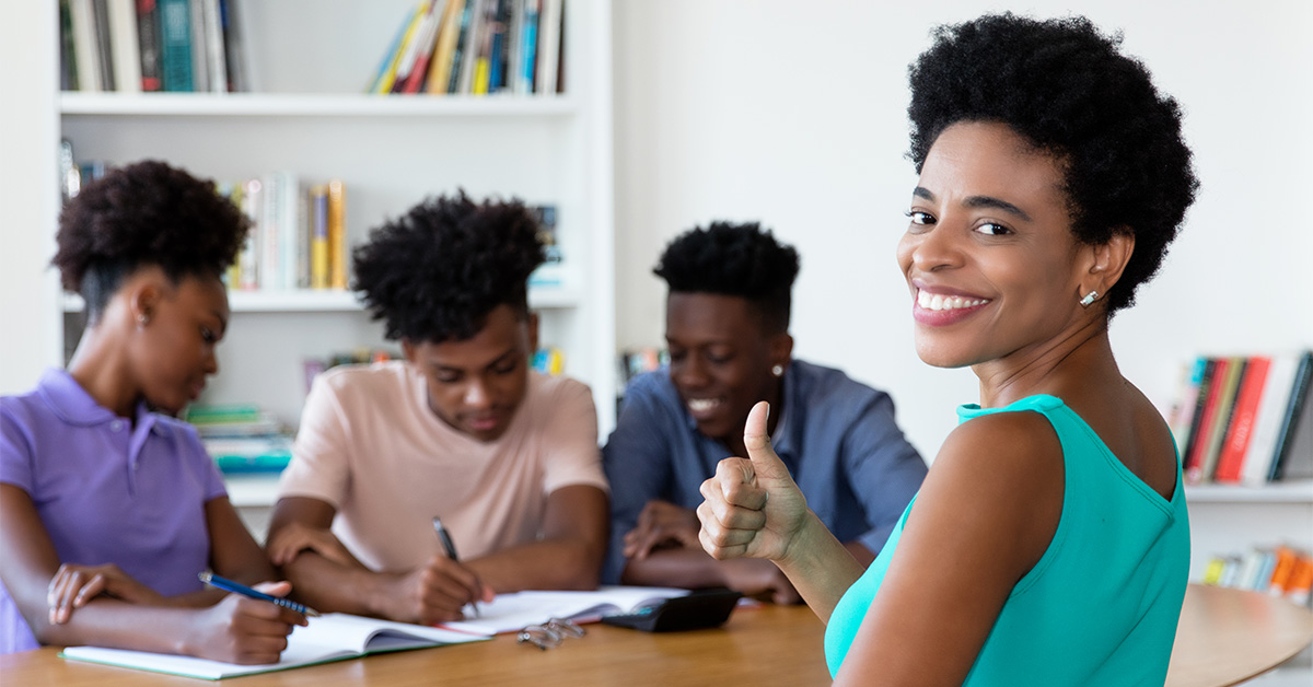 Imagem mostra quatro jovens negros reunidos em uma mesa. Três deles estão de um lado, observando algo que está sendo escrito por um deles em um caderno. A outra jovem, que pode ser a professora dos demais, está à frente, fazendo sinal de positivo e sorrindo para a foto.