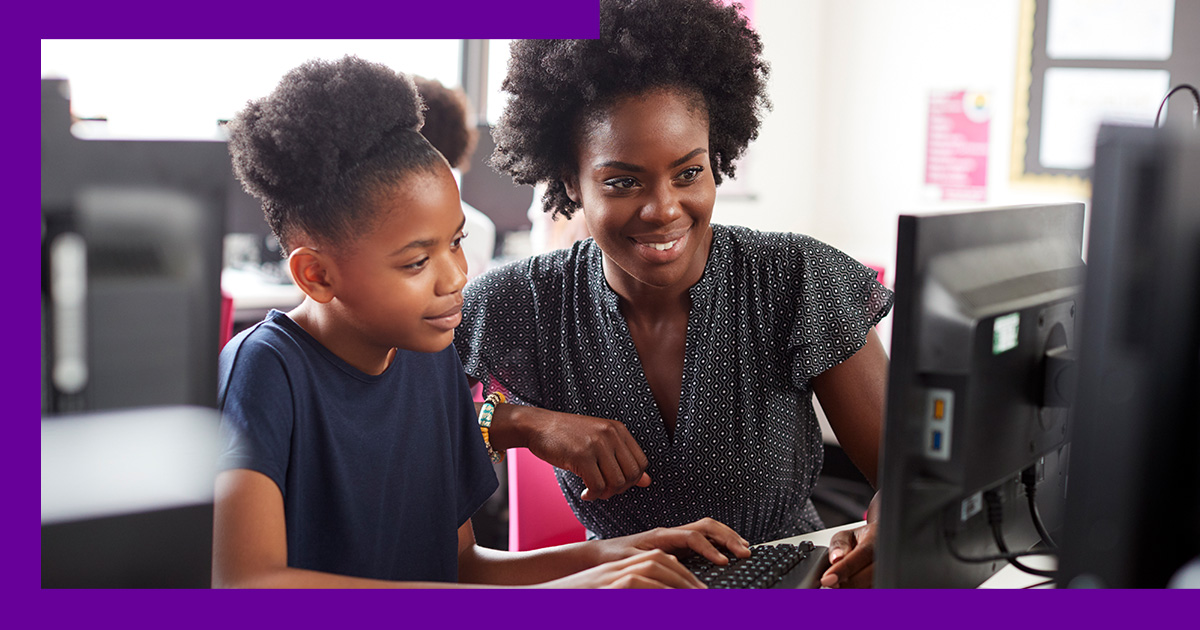 Imagem mostra uma educadora negra ao lado de uma aluna negra. A menina está utilizando um computador e a professora está auxiliando.