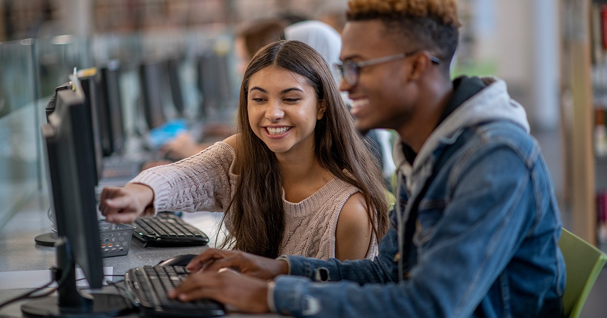 Imagem mostra uma jovem branca e um jovem negro em uma sala de aula com computadores. Os dois estão juntos; ela sorri e aponta para algo na tela do computador, enquanto ele também sorri e observa a tela do aparelho.