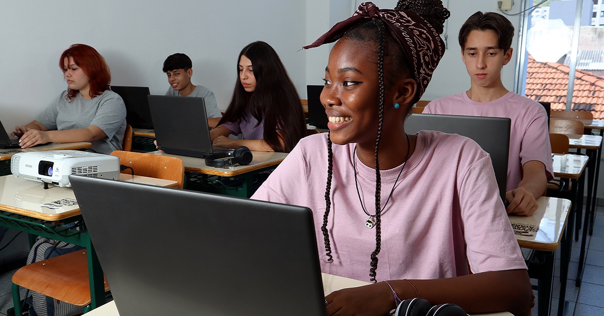 Imagem mostra uma sala de aula onde há seis estudantes. Todos estão sentados em carteiras, utilizando notebooks. Em primeiro plano, há uma jovem negra, que está sorrindo.