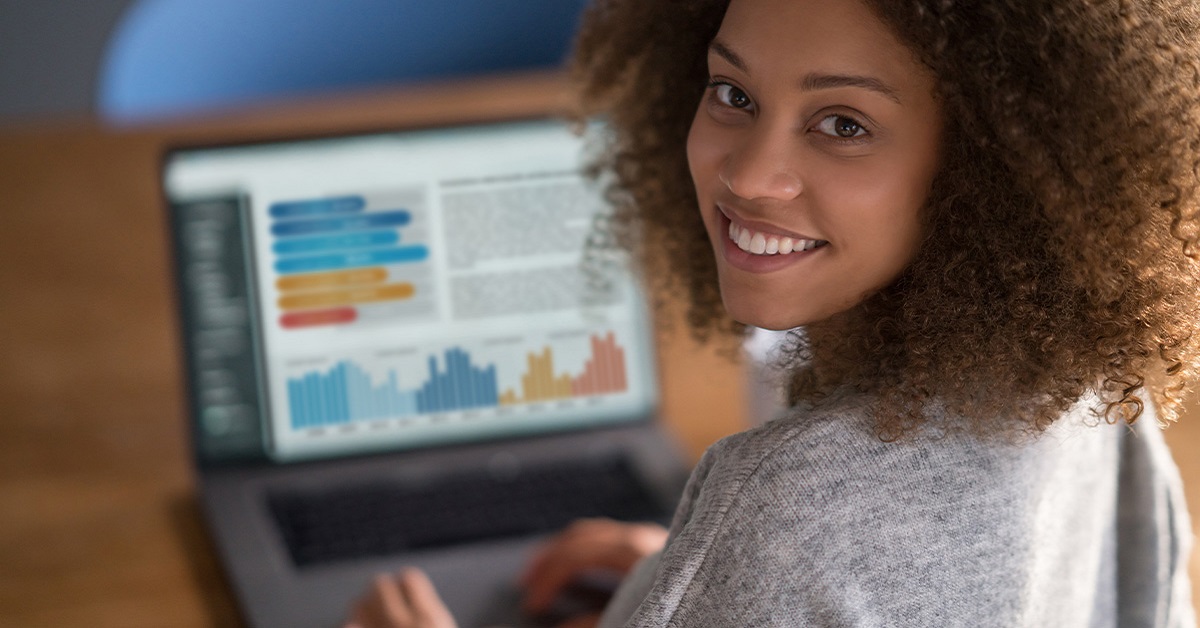 Imagem mostra uma jovem negra olhando para a foto e sorrindo. Ao fundo, é possível ver um computador que está sendo usado pela jovem. Na tela, há alguns gráficos.