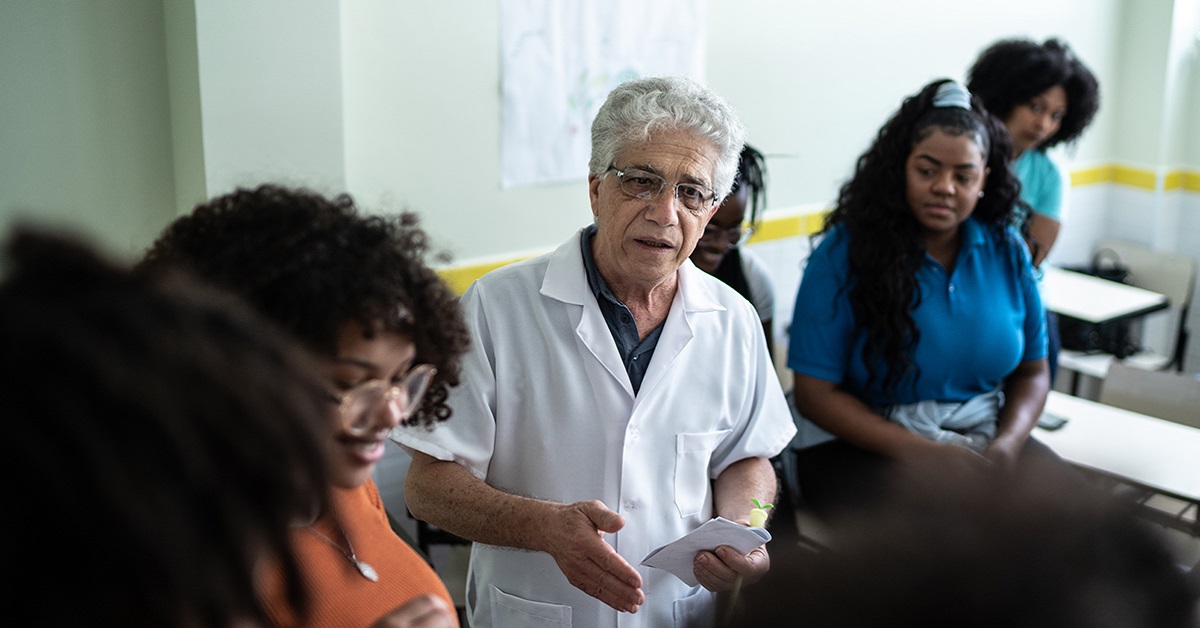 Imagem mostra um professor no centro da imagem. Ele está rodeado por cinco estudantes. Além disso, segura um papel com uma das mãos. Ele parece estar falando algo para os alunos. Todos estão em uma sala de aula.