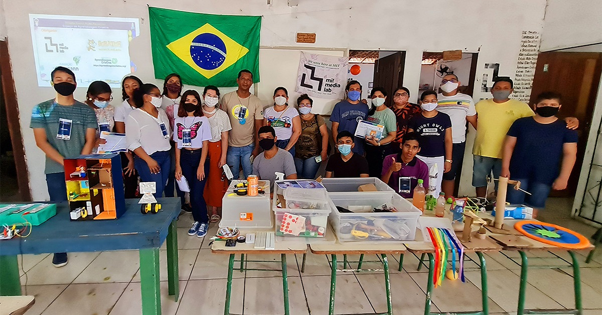 Imagem mostra um grupo de 20 pessoas reunidos em um ambiente onde há mesas e objetos pedagógicos sobre elas. Ao fundo, uma bandeira do Brasil e uma imagem de projetor refletida na parede. O grupo é formado por homens, mulheres e jovens.