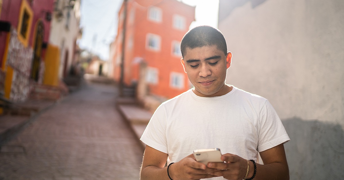 Jovem está mexendo em um aparelho de celular enquanto caminha por uma rua dentro de uma comunidade na América Latina. Ele é moreno, tem os cabelos curtos e usa uma camiseta branca.