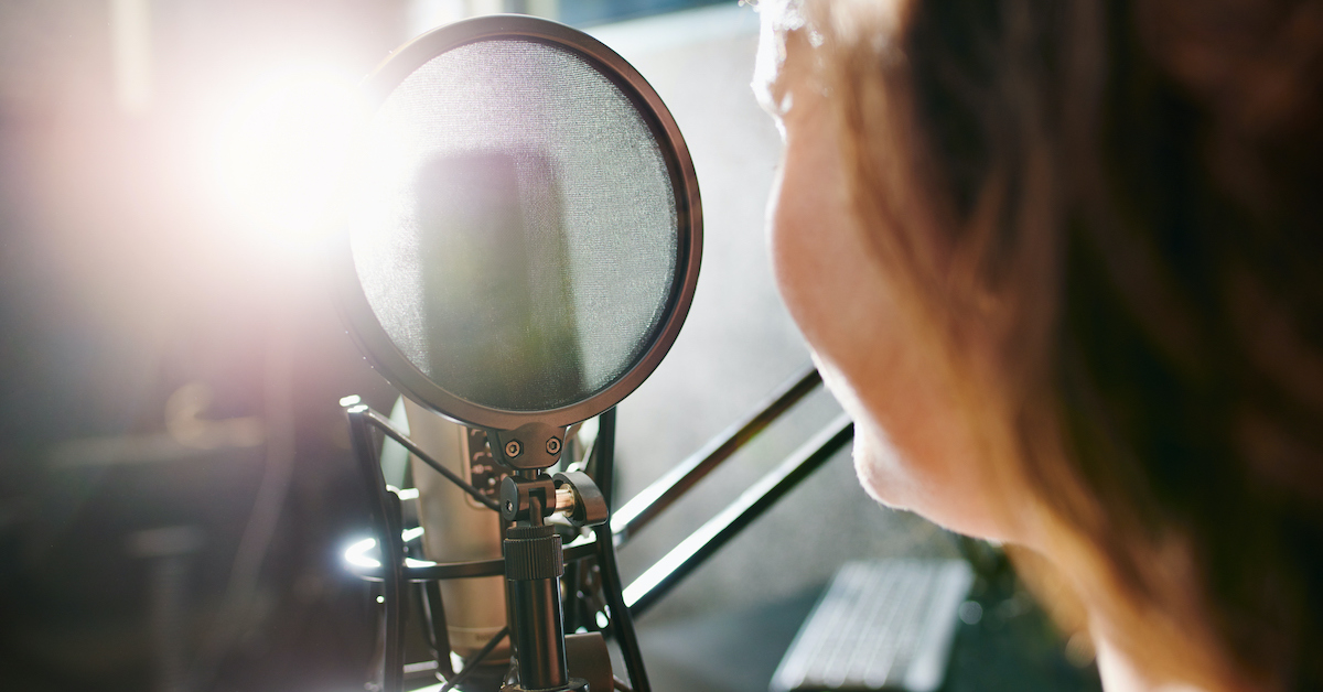 Mulher de costas, falando ao microfone, gravando um áudio, em ambiente escolar.