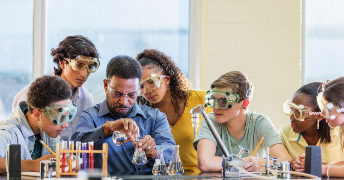 Imagem mostra professor e alunos reunidos debatendo e, ao mesmo tempo, realizando um experimento em laboratório.