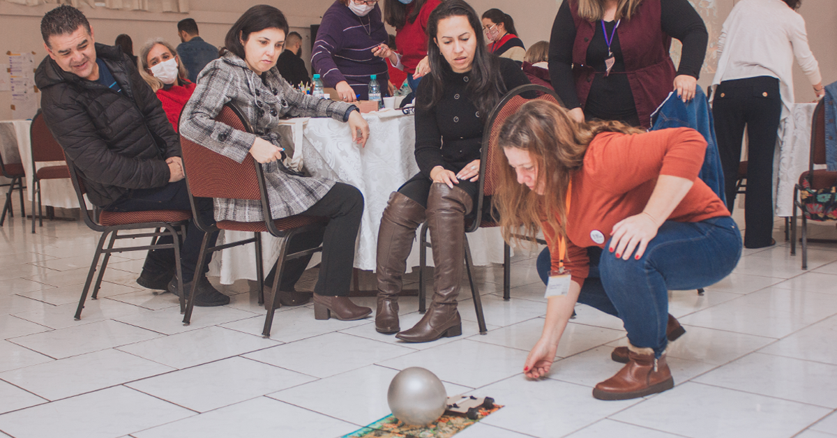 Imagem mostra um grupo de professores (homens e mulheres) participando de uma dinâmica durante o evento. Uma professora está interagindo com uma bola no chão, enquanto os demais professores observam sua ação, alguns professores estão sentados e outros estão em pé.