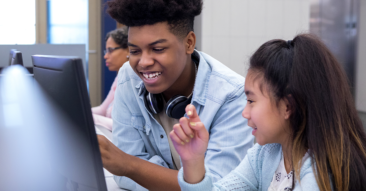 Imagem mostra um adolescente negro e uma adolescente com traços orientais juntos olhando para a tela de um computador. Ao fundo, há uma mulher negra usando um computador