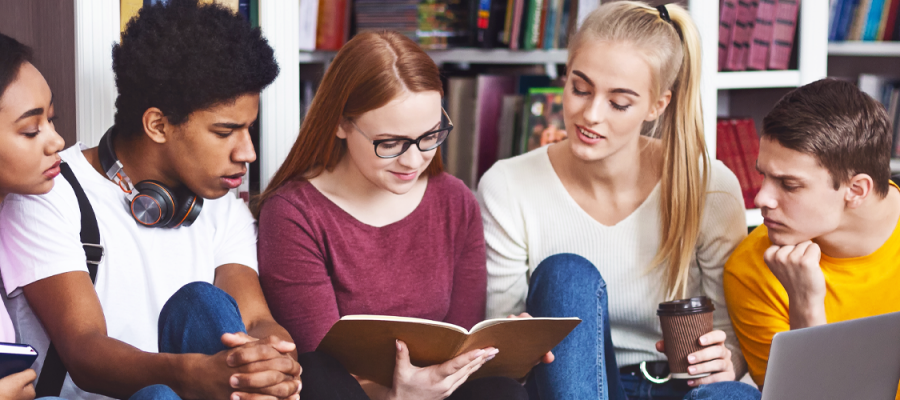 Imagem mostra cinco jovens estudantes reunidos. São dois meninos, sendo um deles negro, e três meninas. Eles estão sentados em frente à uma estante com livros. Dois jovens estão com livros nas mãos. Há um notebook sendo usado por eles. (Itinerário Áreas do Conhecimento)