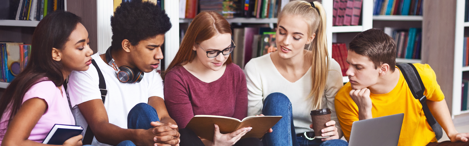 Imagem mostra cinco jovens estudantes reunidos. São dois meninos, sendo um deles negro, e três meninas. Eles estão sentados em frente à uma estante com livros. Dois jovens estão com livros nas mãos. Há um notebook sendo usado por eles. (Itinerário Áreas do Conhecimento)